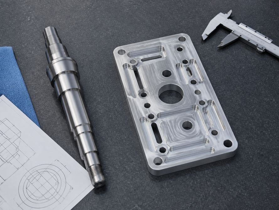 Cylindrical turned shaft beside a milled aluminum plate with pockets and slots